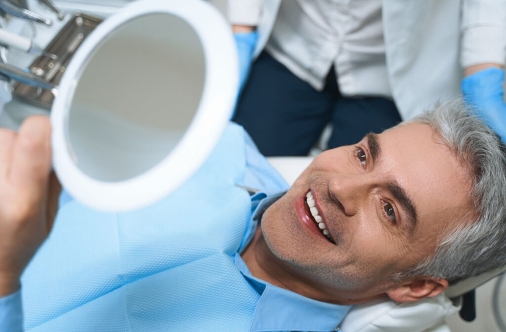 A man sitting in a dentist's chair, holding up a mirror and smiling with his dentist in the background