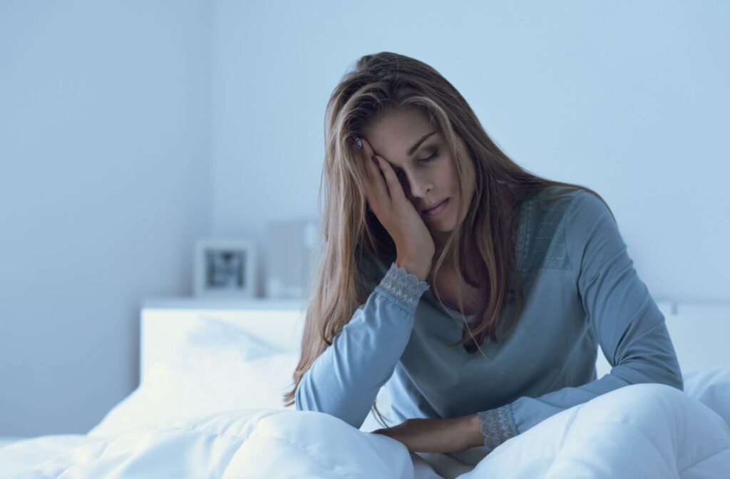 A woman in blue in a blue-tinged room sitting up in bed and holding her right hand to her face with her eyes closed