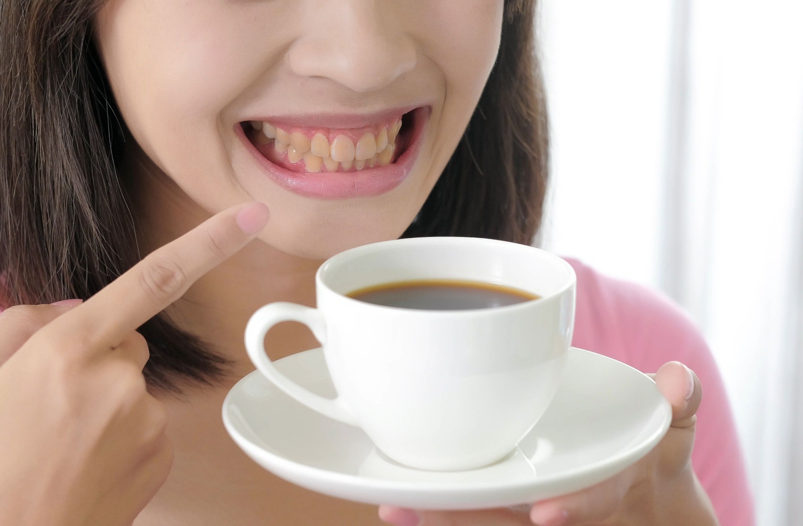 Smiling woman pointing at her yellow teeth while holding a cup of coffee.
