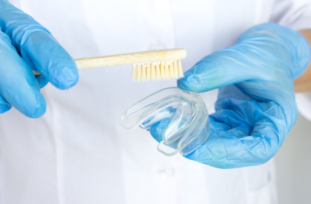 Dentist wearing blue surgical gloves holding a transparent mouth guard with a bamboo toothbrush in a clinical setting