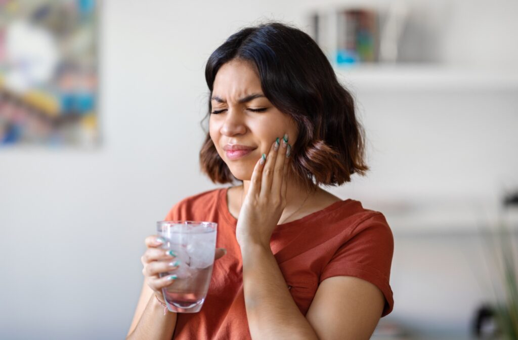 A woman holding a glass of ice water in her right hand and holding her left jaw with her left hand from tooth sensitivity.