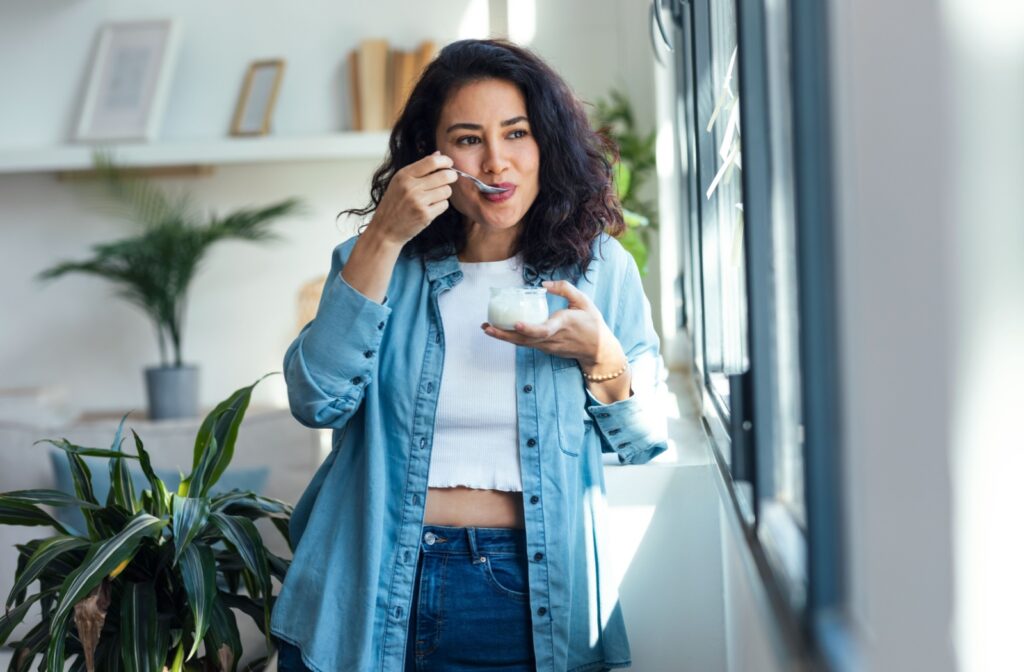Woman standing by a window, eating yogurt with a spoon, dressed in a denim shirt and white top enjoying yogurt after dental implant surgery.