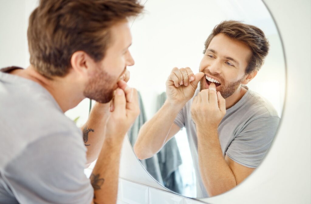 Man in standing at a bathroom mirror, carefully flossing his teeth after dental implant surgery. 
